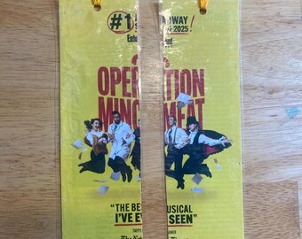 May include: Two yellow rectangular bookmarks featuring the Operation Mincemeat musical promotional art. Each bookmark has a yellow tassel at the top, text reading #1 Broadway 2025, and images of performers in mid-air with scattered papers.