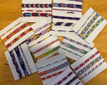 May include: A collection of handmade woven friendship bracelets featuring various geometric patterns and vibrant color combinations, including red, blue, yellow, purple, pink, and green, each mounted on individual white rectangular display cards.