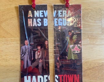May include: Two rectangular paper bookmarks for the musical Hadestown, each encased in clear plastic with a red string tassel at the top. The bookmarks feature promotional imagery, cast photos, and the text A NEW ERA HAS BEGUN and HADESTOWN.