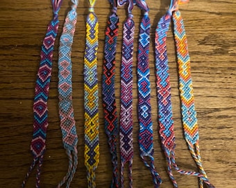 May include: Seven handmade woven friendship bracelets featuring intricate geometric patterns in various color combinations including blue, purple, yellow, orange, red, and pink. Each bracelet has a looped end and braided ties at the bottom.