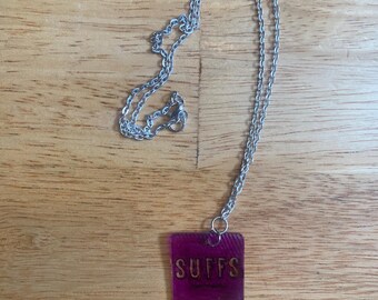 May include: A silver-toned metal chain necklace featuring a square, translucent purple pendant. The pendant has a textured, ribbed surface and displays the word SUFFS in bold, dark lettering centered on the front face of the square piece.