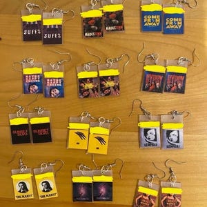 May include: A collection of handmade earrings featuring various designs. Each earring pair has a rectangular shape with a yellow top and a printed image. The images include text like "SUFFS", "HADESTOWN", and "COME FROM AWAY".