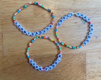 May include: Three circular bracelets made of multicolored seed beads. Each bracelet features white plastic letter beads. One bracelet spells SCHMIGADOON, another spells CROSS THAT BRIDGE, and the third bracelet spells CORN PUDDIN in black capital letters.