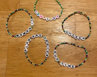 May include: Five circular beaded bracelets made of small multicolored glass beads. Each bracelet features white plastic letter beads spelling out the phrases: LEMPICKA, WOMAN IS, PLAN AND DESIGN, UNSEEN, and OUR TIME. The beads are arranged in a single strand.