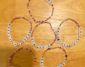 May include: Five circular beaded bracelets made of small red, white, and blue beads. Each bracelet features white letter beads spelling out phrases: RAGTIME, WHEELS OF A DREAM, NEW MUSIC, MAKE THEM HEAR YOU, JOURNEY ON, and BACK TO BEFORE.