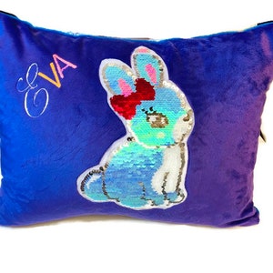 May include: A blue plush pillow with a sequin bunny design. The bunny has a red bow and the pillow has the name "EVA" embroidered on it.