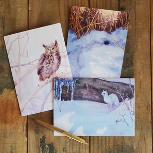 May include: Three watercolor greeting cards featuring winter scenes. The first card shows an owl perched on a branch in a snowy forest. The second card shows a snowy landscape with a log and icicles. The third card shows a weasel walking on snow.