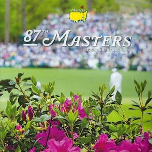May include: The cover of the 87th Masters Tournament Journal, featuring a green golf course with a crowd of spectators in the background. The journal cover also features a large pink azalea bush in the foreground and the text "87th Masters Tournament Journal" and "April 3-9, 2023" in white.