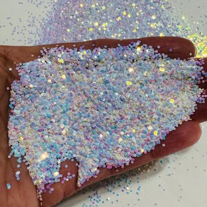 Op de afbeelding: Een close-up van iriserende witte en blauwe glitter met een holografische afwerking. De glitter is verspreid over een wit oppervlak en een hand houdt een hoop glitter vast.