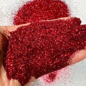 May include: A close-up of a hand holding a pile of fine red glitter. The glitter is sparkling and has a metallic sheen.