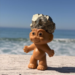 May include: A vintage troll doll with a brown body and a large, textured, metallic pyrite stone in place of hair. The doll stands on a wooden surface with the ocean and blue sky in the background.