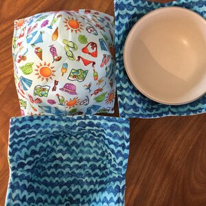 May include: Three fabric bowl cozies. One is blue with a wave pattern, one is blue with a beach-themed pattern, and one is blue with a wave pattern and has a brown bowl inside.