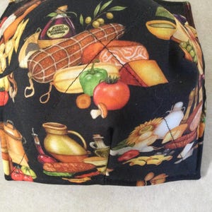 May include: A black fabric item with a colorful print of Italian food items. The design includes a salami, cheese, tomatoes, peppers, and a bottle of balsamic vinegar. The item is square-shaped.