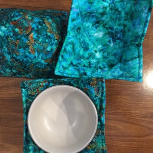 May include: A set of teal bowl cozies and a matching square hot pad. The fabric features a pattern of sea turtles in shades of orange and gold. The bowl cozy is designed to hold a bowl, and the hot pad is square.