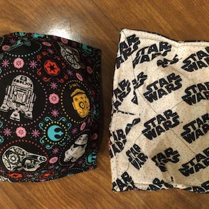May include: Two fabric squares with different Star Wars themed patterns. One square has a black background with colorful Star Wars characters and symbols. The other square has a white background with repeated black "Star Wars" text.