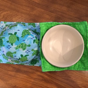 May include: A set of two fabric bowl cozies. One is blue with a sea turtle pattern and the other is green. Both cozies are quilted and have a white bowl inside.