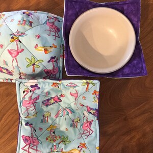 May include: Three square bowl cozies with a flamingo and tropical theme. The cozies are made of a light blue fabric with pink flamingos, palm trees, and umbrellas. The cozies have a purple border.