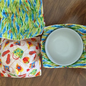 May include: Three quilted fabric bowl cozies. One is green and blue with a wavy pattern. Another is white with a red, yellow, and blue pattern of beach-themed items. The third is green and blue with a wavy pattern and has a white bowl inside.