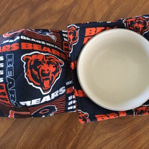 May include: Two blue and orange fabric pot holders with the Chicago Bears logo and the word "Bears" repeated. A white ceramic bowl is sitting on top of one of the pot holders.