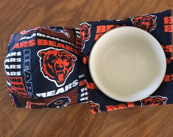 May include: Two blue and orange fabric pot holders with the Chicago Bears logo and the word "Bears" repeated. A white ceramic bowl is sitting on top of one of the pot holders.
