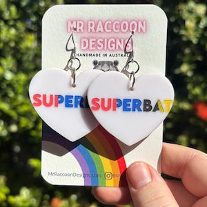 May include: White heart-shaped earrings with the word "SUPER SUPERBAT" in red, blue, and yellow. The earrings are on a white card with the text "MR RACCOON DESIGN" and a rainbow design.