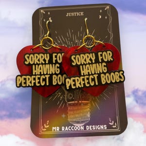 May include: Heart-shaped earrings with the text "SORRY FOR HAVING PERFECT BOOBS" in gold. The red earrings have a textured design and gold ear wires. They are displayed on a black card with "JUSTICE" at the top. The earrings are a fun accessory.