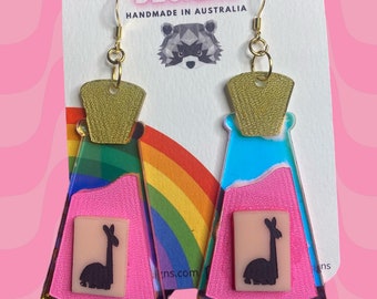 May include: A pair of gold and iridescent earrings shaped like potion bottles with a pink and blue liquid inside. Each bottle has a small square with a black silhouette of a llama inside. The earrings are handmade in Australia.