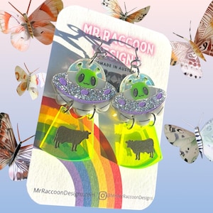 Op de afbeelding: Nieuwe oorbellen met een UFO-ontwerp, met een groene alien en een koeienafbeelding. De oorbellen zijn gemaakt van acryl en hebben een zilveren glitterrand. De achtergrond bevat een regenboog en vlinders.