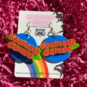 May include: A pair of novelty earrings featuring a blue planet design with a green ring and the words "Coolmath Games" in red and yellow. The earrings are suspended from gold-colored hooks. The background includes a rainbow and the text "Mr Raccoon Designs".