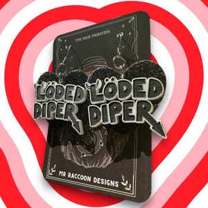 May include: Black glitter heart-shaped earrings with the words "LÖDED DİPER" in a bold, outlined font. The earrings hang from a black card with the text "THE HIGH PRIESTESS" and "MR RACCOON DESIGNS" on a red and pink striped background.