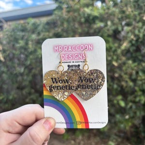 May include: Heart-shaped earrings with gold glitter and the words "Wow, genetic" in black. The earrings hang from gold-colored hooks and are attached to a white card with the text "MR RACCOON DESIGNS" and a rainbow.