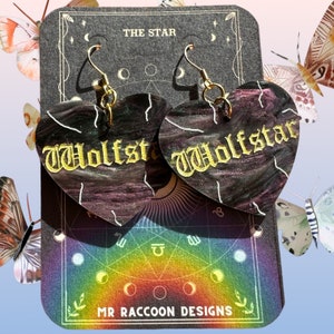 May include: A pair of heart-shaped earrings with a black and purple marbled finish. The earrings feature the word "Wolfstar" written in gold script. The earrings are displayed on a black card with a rainbow design and the text "MR RACCOON DESIGNS".