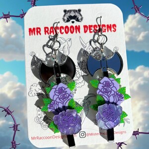 Op de afbeelding: Hangende oorbellen met een zwart bijlontwerp, paarse rozen en groene bladeren. De oorbellen worden getoond op een witte kaart met de tekst "MR RACCOON DESIGNS". De achtergrond toont een blauwe lucht met wolken.