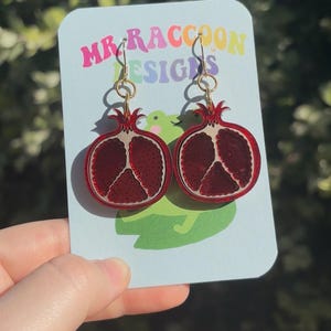 May include: A pair of red pomegranate earrings with gold-colored hooks. The earrings are on a light blue card with the text "MR RACCOON DESIGNS" in colorful letters. The earrings are a fruit-themed accessory.