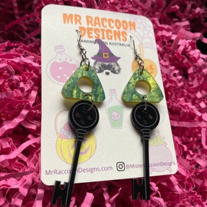 Op de afbeelding: Hangende oorbellen met een zwart sleutelontwerp dat aan een groen en gele driehoek hangt. De oorbellen worden gepresenteerd op een kaart met de tekst "MR RACCOON DESIGNS HANDMADE IN AUSTRALIA". De achtergrond is roze.