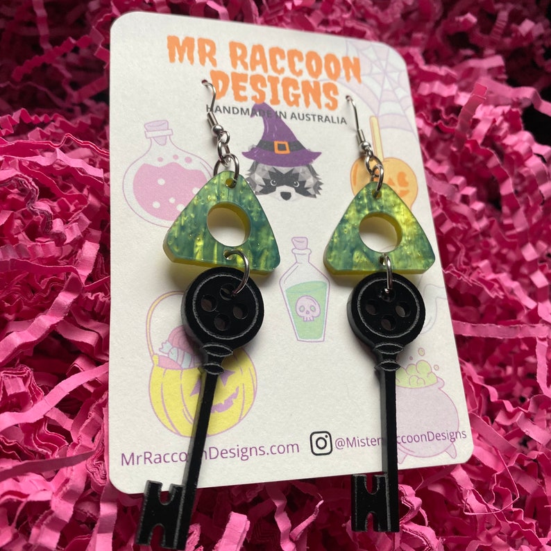 Op de afbeelding: Hangende oorbellen met een zwart sleutelontwerp dat aan een groen en gele driehoek hangt. De oorbellen worden gepresenteerd op een kaart met de tekst "MR RACCOON DESIGNS HANDMADE IN AUSTRALIA". De achtergrond is roze.