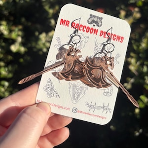 Op de afbeelding: Koperkleurige oorbellen in de vorm van bijlen, met ingewikkelde schildontwerpen. De oorbellen worden gepresenteerd op een witte kaart met de tekst "MR RACCOON DESIGNS" en andere illustraties. De achtergrond is wazig groen gebladerte.