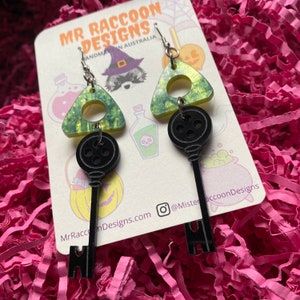 Op de afbeelding: Een paar Halloween-thema oorbellen. Elke oorbel heeft een zwarte sleutelhanger die onder een groen en gele driehoekige bedel hangt. De oorbellen worden gepresenteerd op een kaart met de tekst "MR RACCOON DESIGNS MADE IN AUSTRALIA".