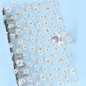 May include: A clear, daisy-patterned binder with silver metal rings and a snap closure. The binder's cover is adorned with white and yellow daisy flowers on a light blue background. The binder is designed for organizing papers.