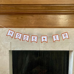 May include: A brown and white banner with the text "HOOK 'EM" hanging above a fireplace. The banner features a white outline of the state of Texas with a brown interior.