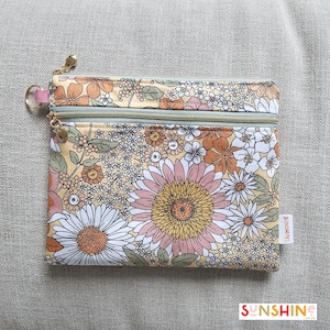 Può includere: Un astuccio rettangolare con stampa floreale e due scomparti con cerniera. Il motivo presenta grandi fiori rosa, bianchi e gialli su sfondo crema. Le cerniere hanno tiretti dorati e un piccolo anello rosa è attaccato al lato. La parola "SUNSHINE" è stampata in basso.