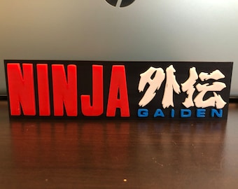 Puede incluir: Un letrero rectangular negro con las palabras "NINJA GAIDEN" en letras rojas y blancas. Los caracteres japoneses para "Gaiden" también están incluidos en blanco.