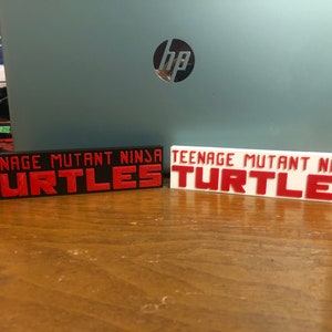 May include: Two rectangular signs with the text "TEENAGE MUTANT NINJA TURTLES" in red. One sign is black, the other is white. The signs are placed on a wooden surface in front of a laptop.