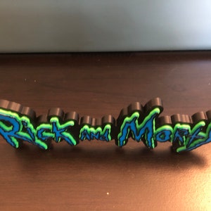 Może przedstawiać: Tr&oacute;jwymiarowy znak z napisem "Rick and Morty" w stylu niebieskiego i zielonego graffiti.