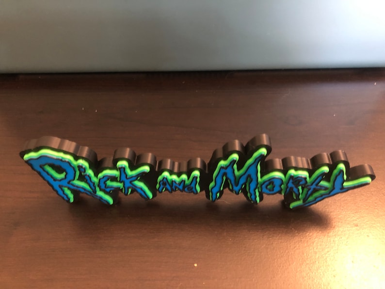 Może przedstawiać: Tr&oacute;jwymiarowy znak z napisem "Rick and Morty" w stylu niebieskiego i zielonego graffiti.