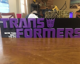 May include: Purple 3D printed sign with the words "Transformers" and the text "More Than Meets The Eye!" on a black background.