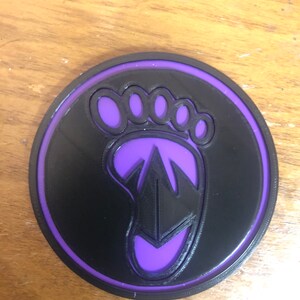 May include: A purple and black circular patch with a footprint design. The footprint has five toes and a stylized arrow inside.
