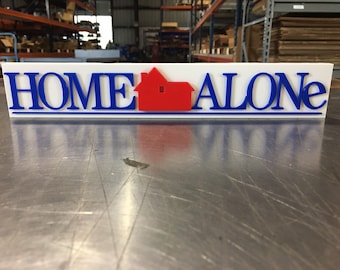 Puede incluir: Señal blanca con las palabras "HOME ALONE" en letras azules y un icono de casa roja en el medio.