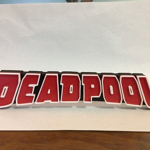 May include: A 3D printed sign that reads "DEADPOOL" in red letters with white outlines on a black background.