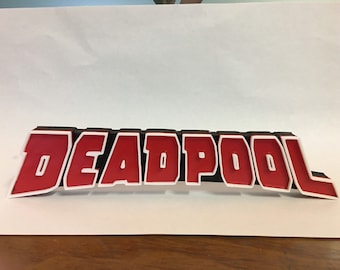 Puede incluir: Un letrero impreso en 3D que dice "DEADPOOL" en letras rojas con contornos blancos sobre un fondo negro.